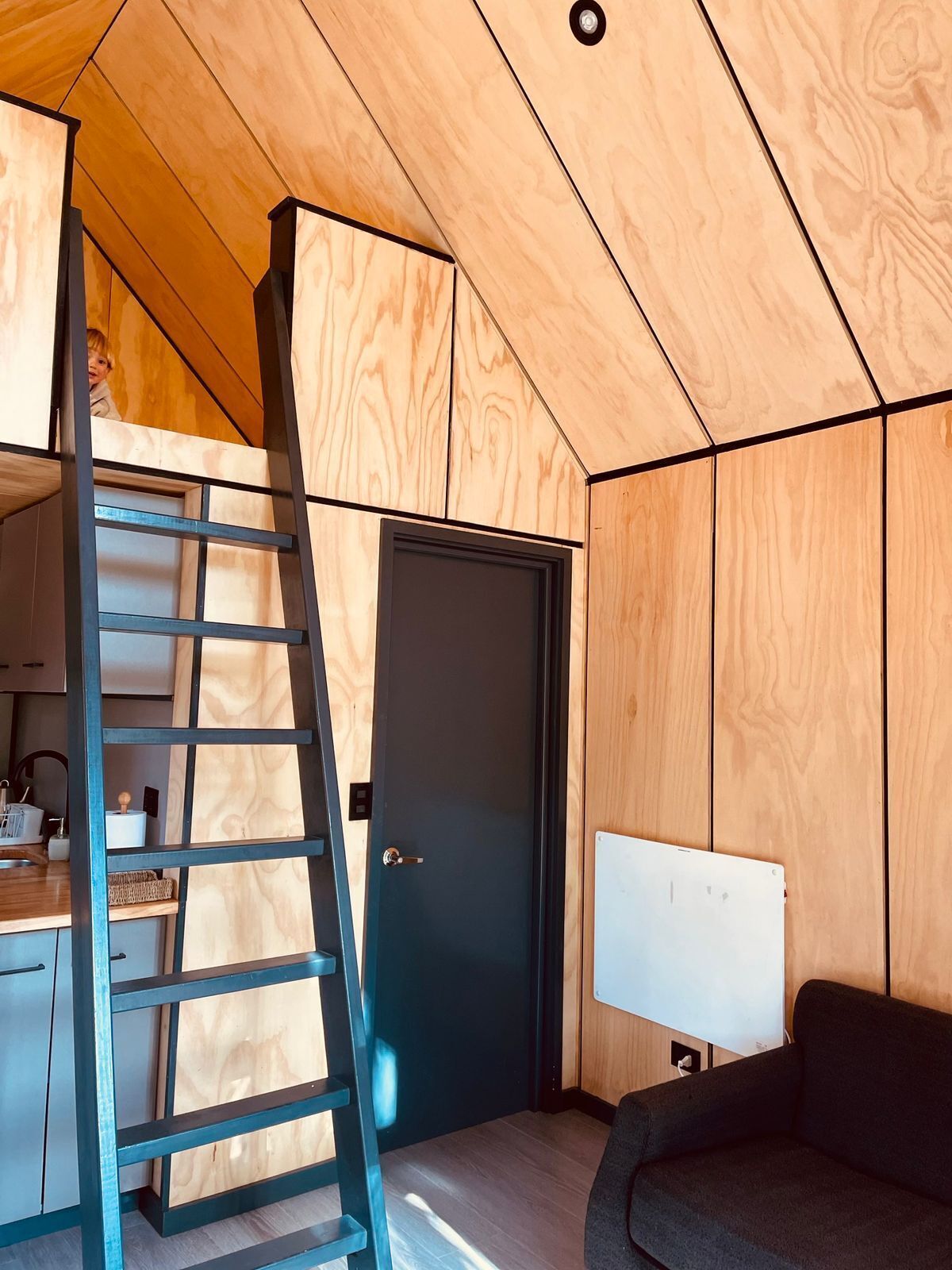 A compact room with a wooden ceiling, a dark door, a whiteboard, a black sofa, and a wooden ladder.