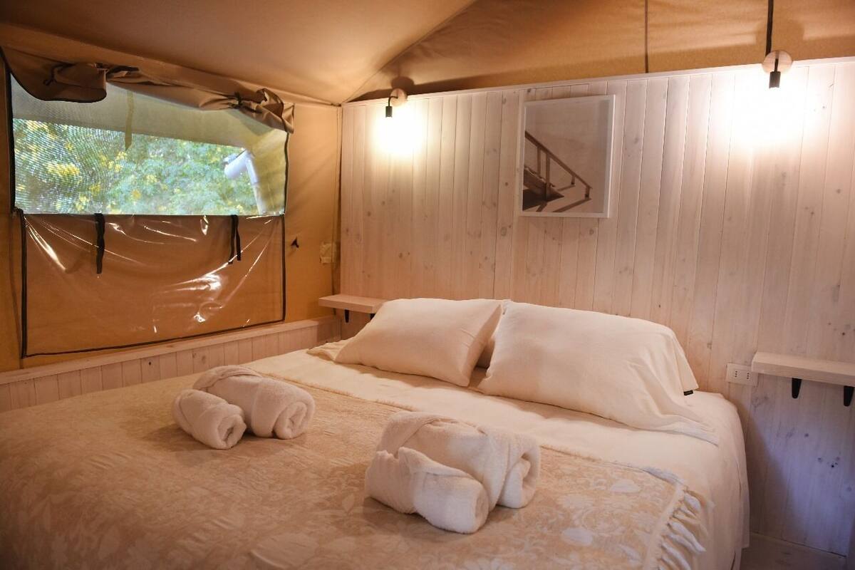 Un dormitorio con paneles de madera, una cama, dos toallas enrolladas, una ventana con vistas y una lámpara fijada a la pared.