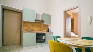 A compact kitchen with a brick backsplash, a stove, and a sink. There is a dining table with chairs and a door leading to another room. - Posillipo Apartment (Napoli)