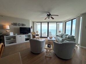 A living room with a television, seating, a ceiling fan, and a view of the ocean.