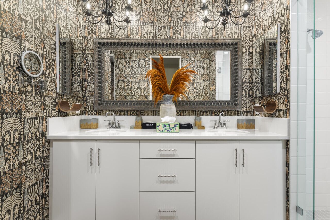 A bathroom with a patterned wallpaper, a large mirror, two sinks, and a vase with dried plants.