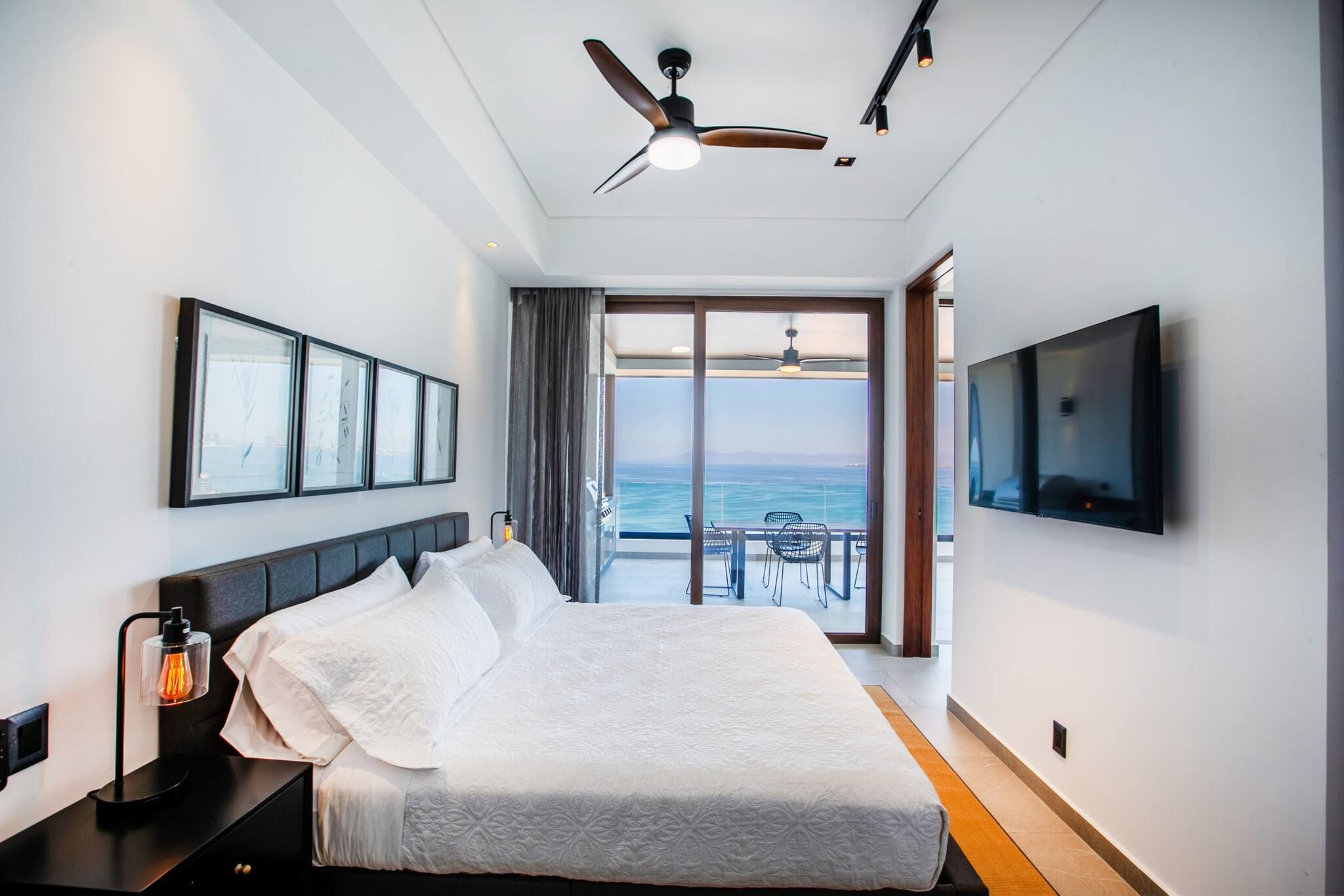 A hotel room with a bed, a TV, a ceiling fan, and a view of the sea.