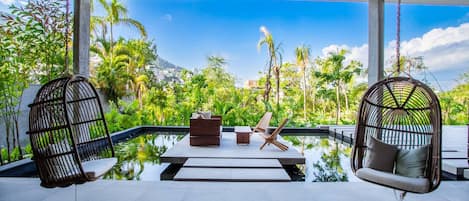 A serene outdoor seating area with a pool, surrounded by lush greenery and a clear blue sky.