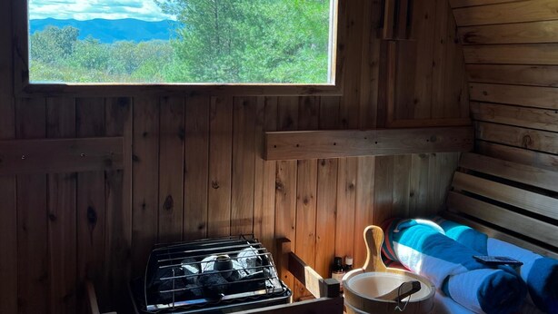 A cabin with a large window overlooking a forest and mountains, a stove, and a bed with a blanket and pillow.