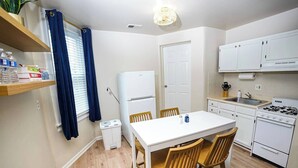 Een kleine keuken met witte kastjes, een fornuis, een spoelbak en een eettafel met stoelen.