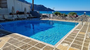 Ein klares, blaues Schwimmbecken mit gefliestem Deck, Liegestühlen und einer Steinmauer mit Blick auf Meer und Berge.