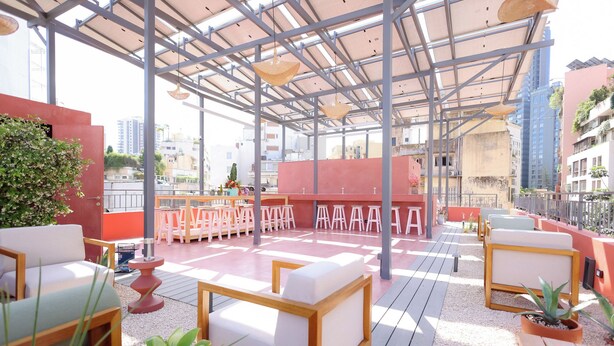 An outdoor seating area with tables and chairs, a red wall, and a view of the cityscape. - Dar Khawla (Beirut)