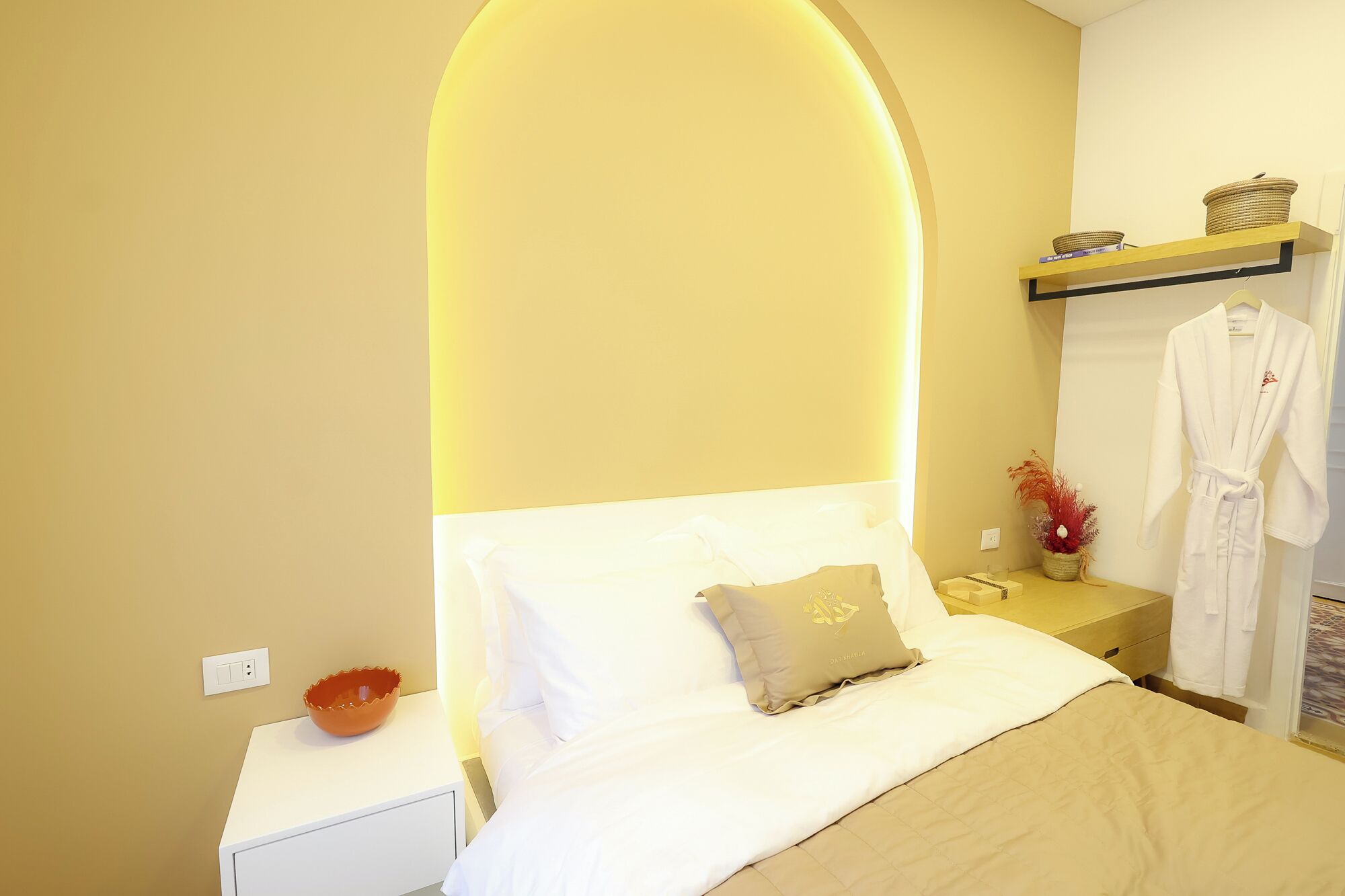 A hotel room with a bed, a nightstand, a robe, and a shelf with books and a basket.