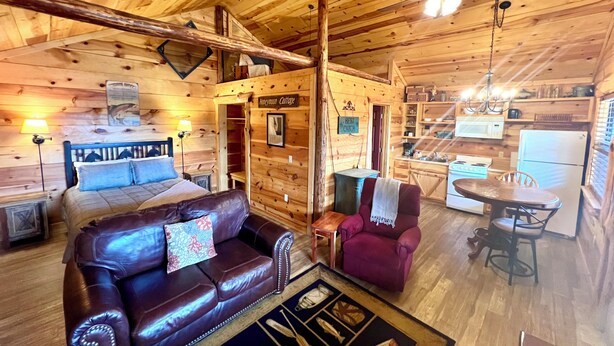 A cozy cabin interior with a wooden ceiling, a bed with a headboard, a leather sofa, a dining table with chairs, and a kitchen area. - Best Catch #49 Cabin in town, walking distance to the courthouse square. (Mountain View)