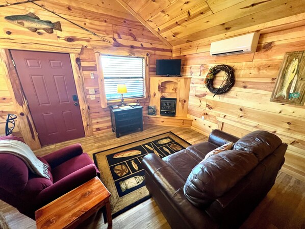 A cozy cabin interior with a wooden ceiling, a fireplace, a television, a plush red sofa, and a wooden coffee table.