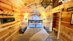 A cozy wooden cabin bedroom with a bed, two bedside lamps, a mirror, and a 'Home' sign on the wall. - Best Catch #49 Cabin in town, walking distance to the courthouse square. (Mountain View)