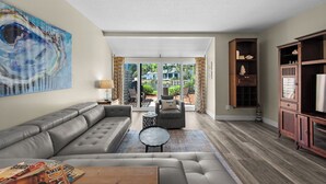 Una sala moderna con un sofá seccional gris, una mesa de centro y un mueble para televisor. Se ve una ventana grande con vistas a una piscina y muebles de exterior.