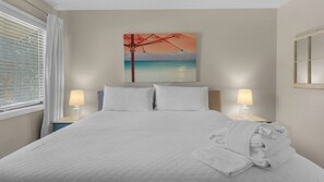 A neatly made bed with white linens, two bedside lamps, a painting of a sunset over the sea, and a window with blinds. - 1/2 Mile to the Beach at Sandestin + Pool: Lake Views at Beachwalk Villas (Miramar Beach)