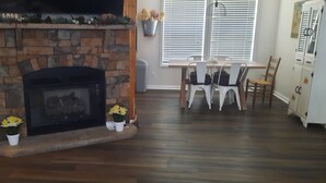 Ein Wohnzimmer mit Kamin aus Stein, Holzboden, einem Esstisch mit Stühlen und einer weißen Kommode.