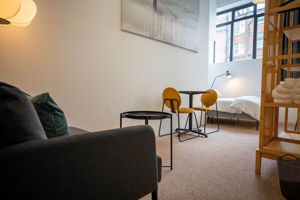 Ein modernes Hotelzimmer mit einer Couch, zwei gelben Sesseln, einem kleinen runden Tisch, einem Bett mit weißer Bettwäsche, einem hölzernen Bücherregal und einem großen Fenster mit Blick auf die Stadt.