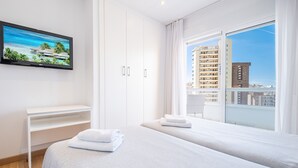Una habitación de hotel con un ventanal grande, un televisor de pantalla plana, una cama con ropa blanca y un armario blanco.