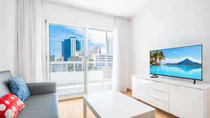 Una sala moderna con un sofá, una mesa de centro, un televisor en un mueble blanco y un ventanal con vista a la ciudad.