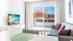 A modern living room with a television displaying a beach scene, a grey sofa with red and black striped pillows, a dining table with green placemats, and a large window with a view of buildings and a clear sky.