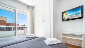 A hotel room with two beds, a TV on the wall, a balcony with a view of the city, and a white desk.