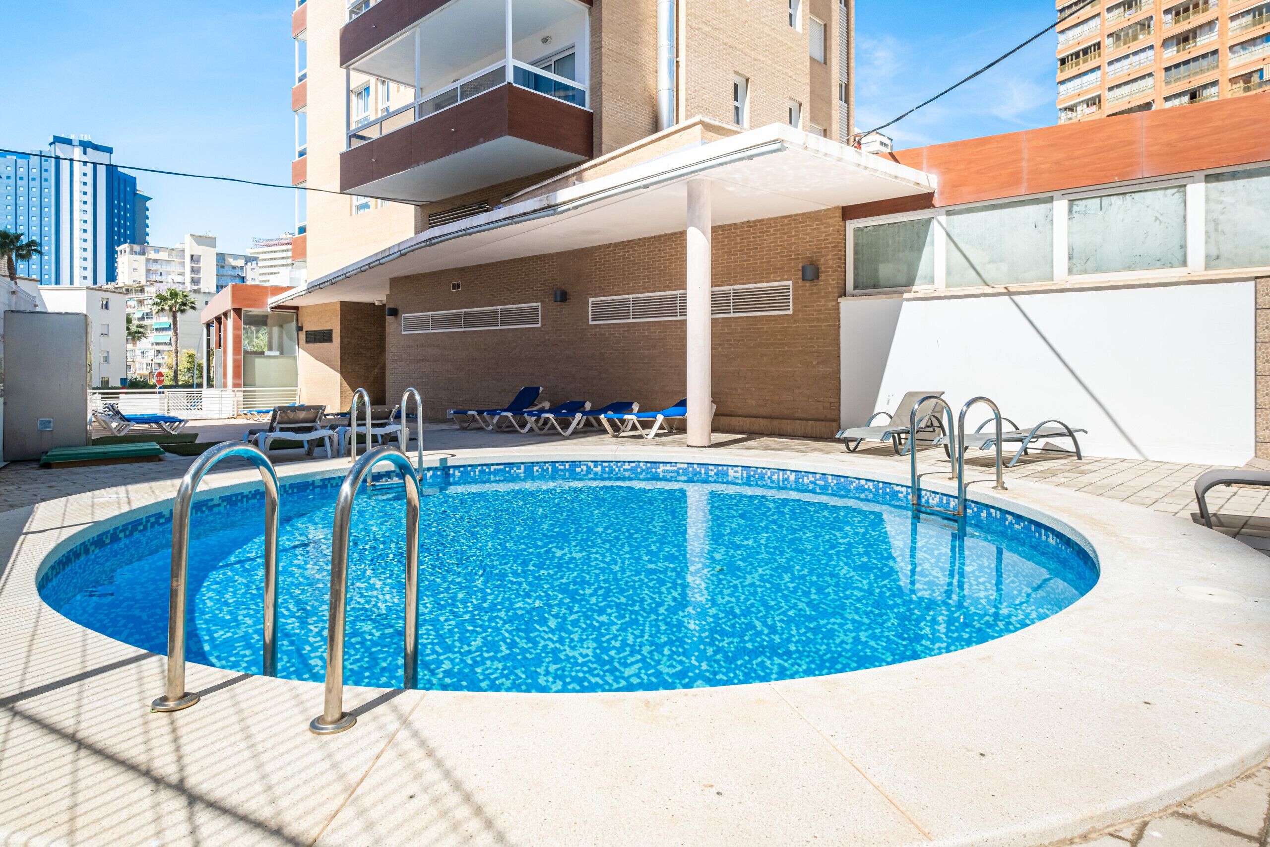 Una piscina con sedie a sdraio e un'area salotto coperta, circondata da edifici.