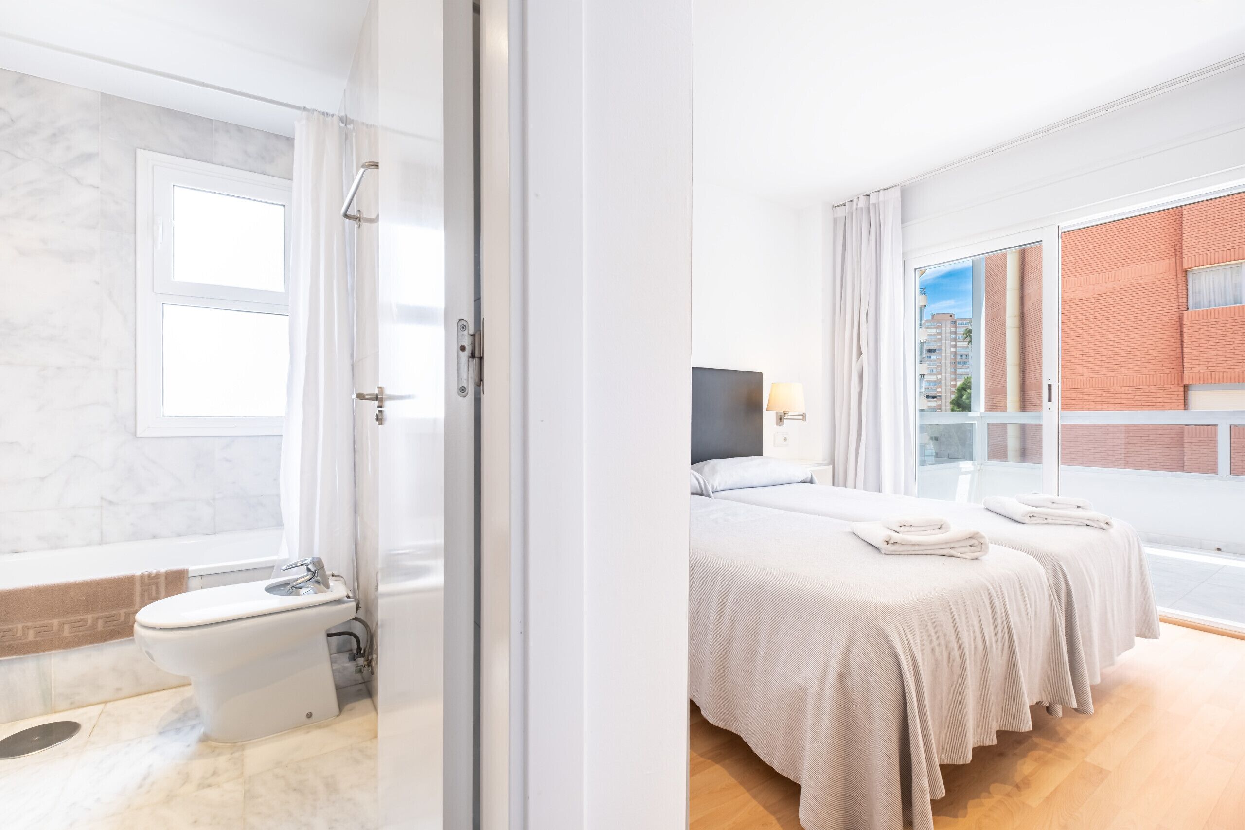 Une salle de bain moderne équipée de toilettes, d’un bidet et d’une douche. Une grande fenêtre offre une vue sur un bâtiment situé à l’extérieur.