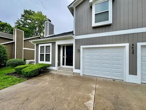 Un immeuble résidentiel à deux étages, doté d’un garage, d’une porte d’entrée et d’un petit jardin devant, agrémenté d’une haie.