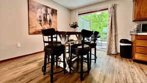 A dining area with a table and four chairs, a painting on the wall, and a view through a window with curtains.
