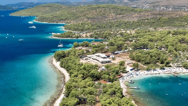 An aerial view of a coastal area with a small island surrounded by clear blue waters, a sandy beach, and dense green vegetation.