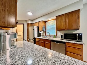Una cocina con encimeras de granito, gabinetes de madera y electrodomésticos de acero inoxidable, además de un ventanal con persianas.