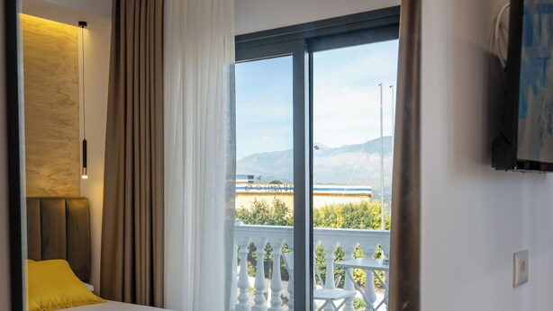 A modern hotel room with a bed, a wooden nightstand, a TV mounted on the wall, and a balcony with outdoor furniture. - Hotel Flori (Sarandë)