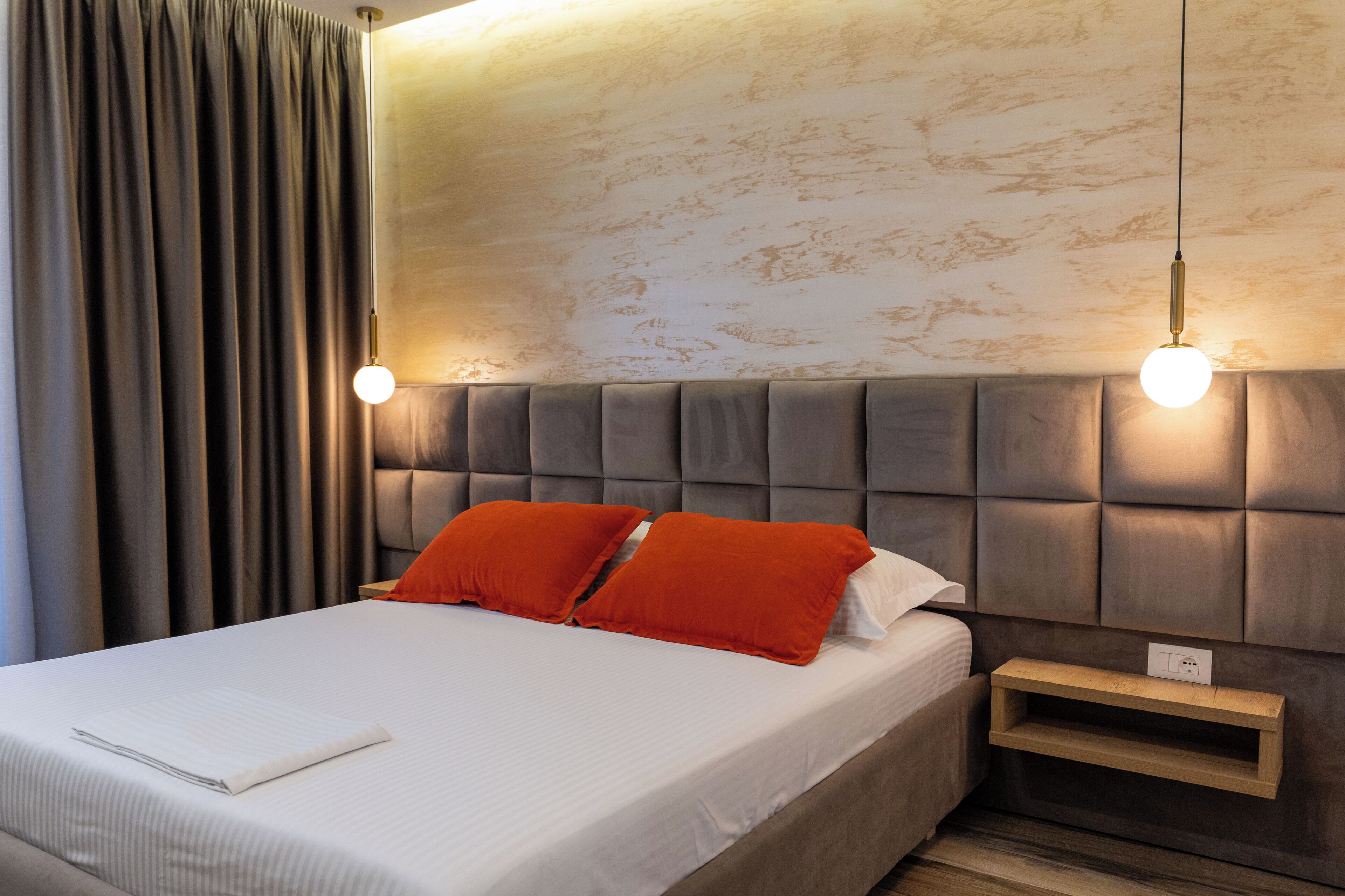 Een net opgemaakt bed met witte lakens en twee oranje kussens, een nachtkastje met een stopcontact, en een hoofdbord met een gestructureerde afwerking.
