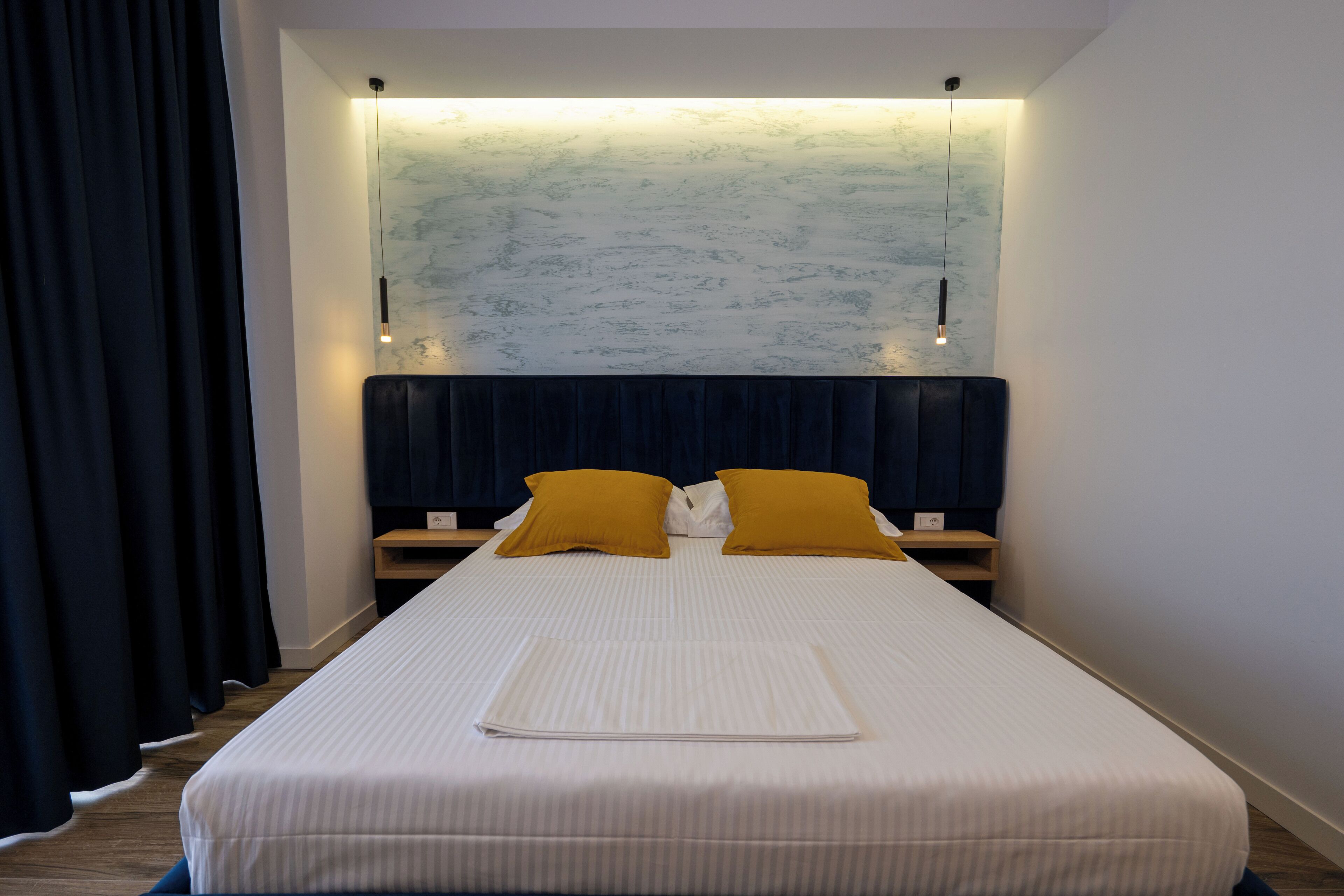 A neatly made bed with white bedding and two mustard pillows, a dark headboard, and a wall-mounted light fixture.