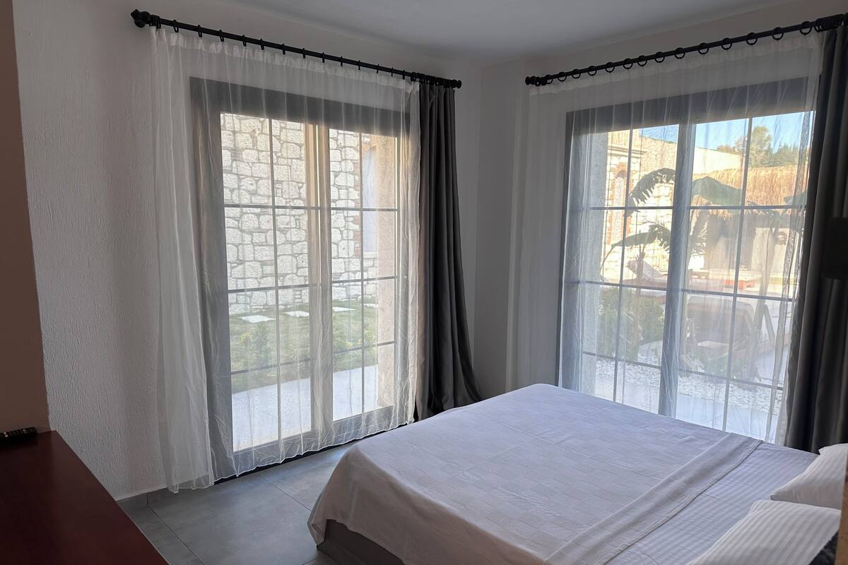 A bedroom with a bed, two windows with curtains, a view of a stone wall, and a tiled floor.