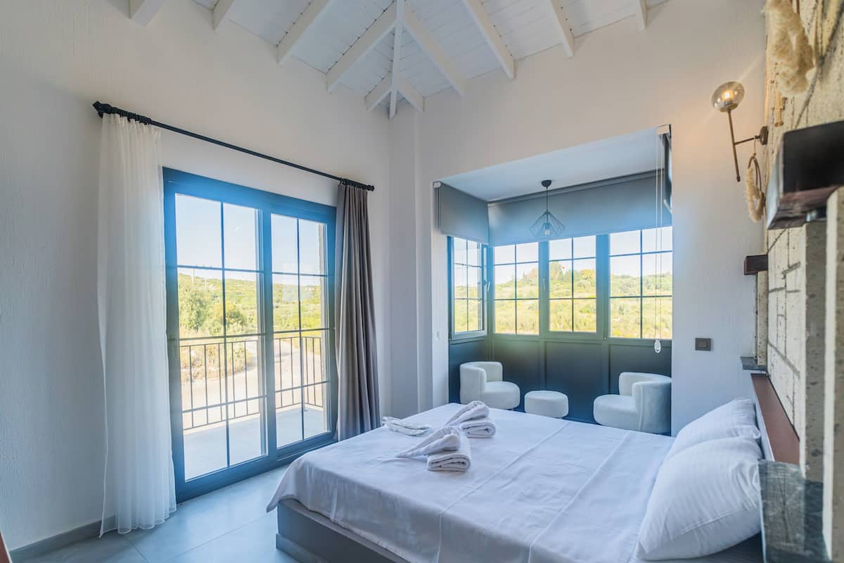 A bedroom with a bed, two chairs, a window with a view of greenery, and a balcony with a railing.