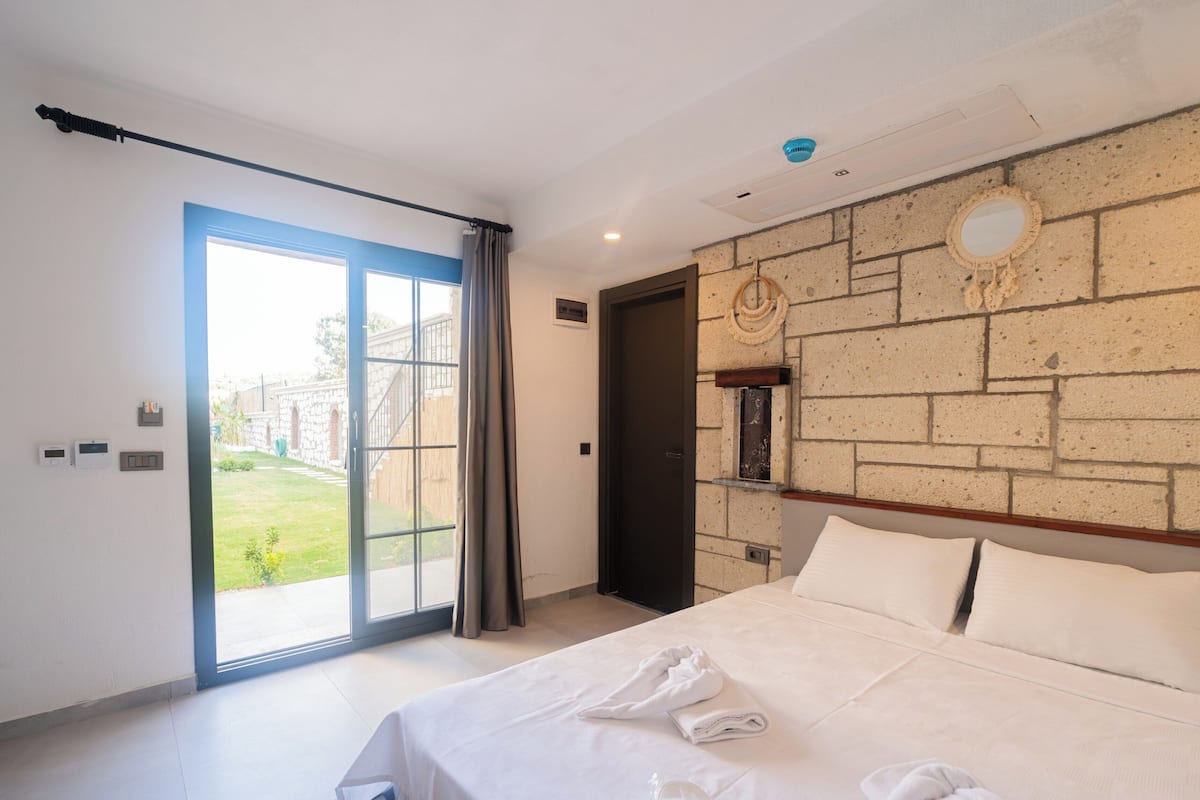 A hotel room with a large bed, a view of the garden through the sliding door, and a stone wall with a mirror.