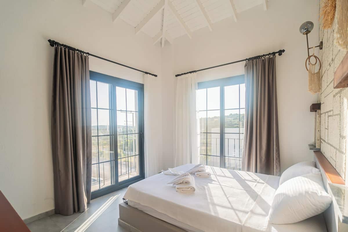 A bedroom with a large bed, two windows with curtains, a view of the outdoors, and a wooden headboard.