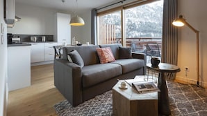 Ein modernes Wohnzimmer mit grauem Sofa, einem Couchtisch und Blick auf eine Küche und einen Balkon.