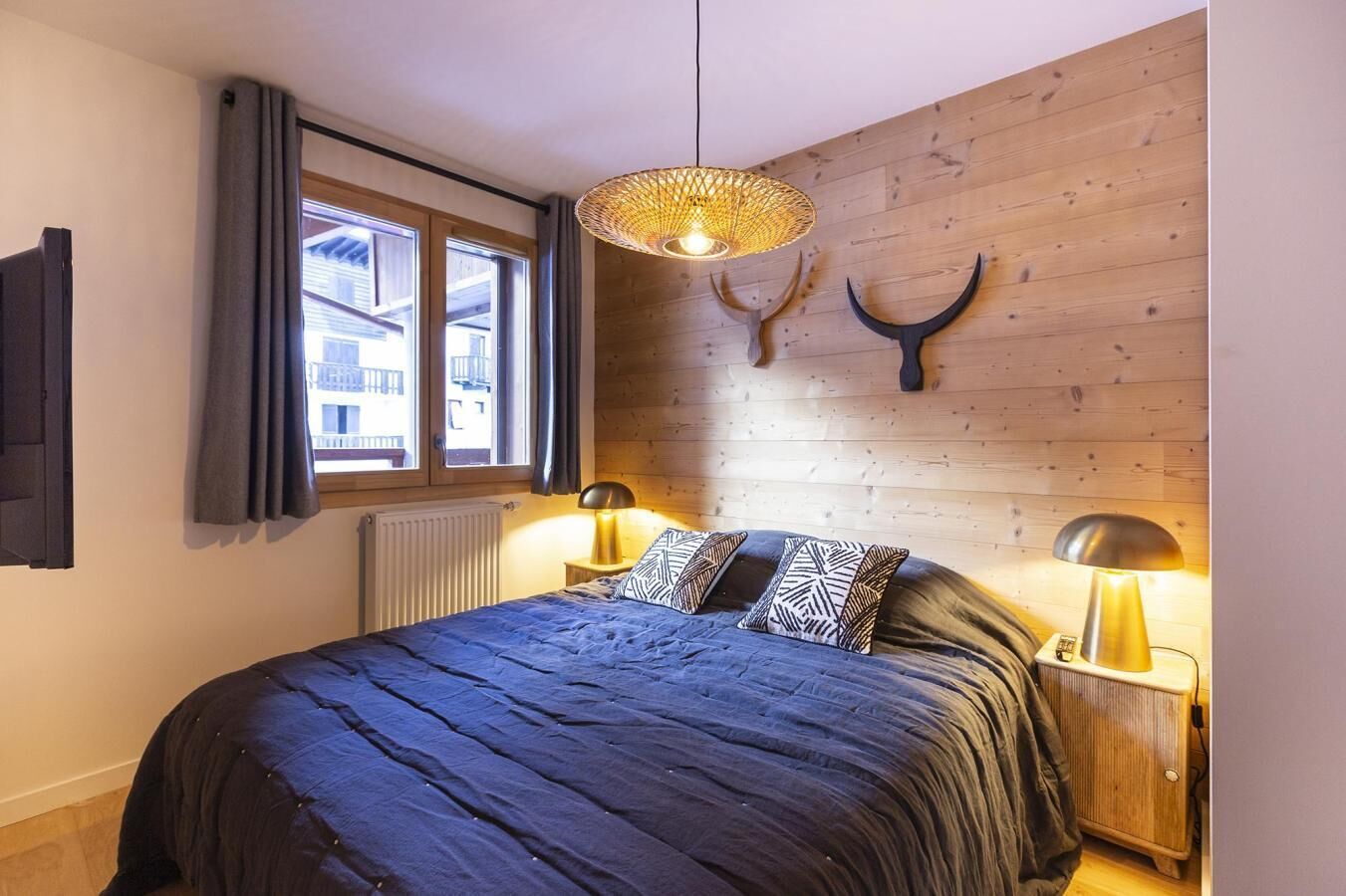 A bedroom with a large bed, two bedside lamps, a window with curtains, a radiator, and a wooden wall with a mounted animal head.