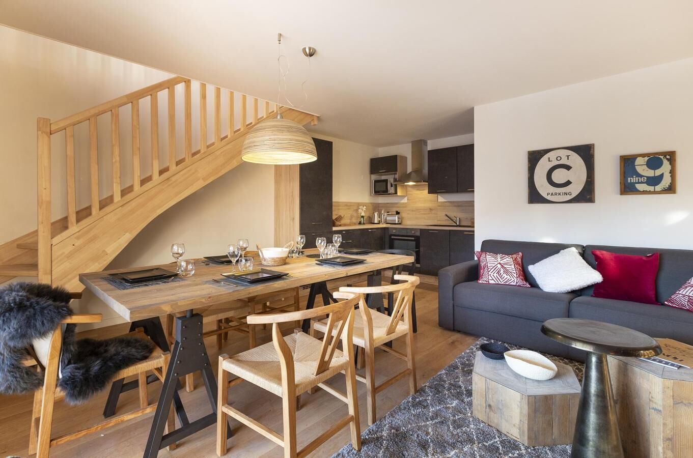 Een moderne woonruimte met een houten trap, een eethoek met een houten tafel en stoelen, een grijze bank met gekleurde kussens, en een keuken op de achtergrond.