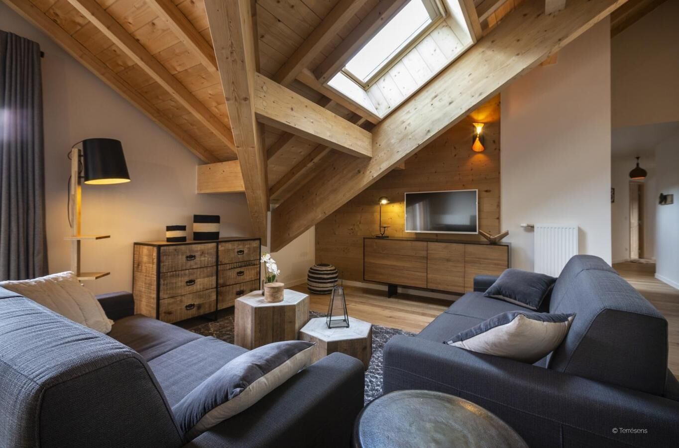 A loft-style living room with a sloped ceiling, wooden beams, a TV, a sofa, and a side table with a lamp.