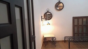 Een kamer met een houten vloer, een tafeltje met een lamp, een spiegel aan de muur, en een raam met jaloezieën.