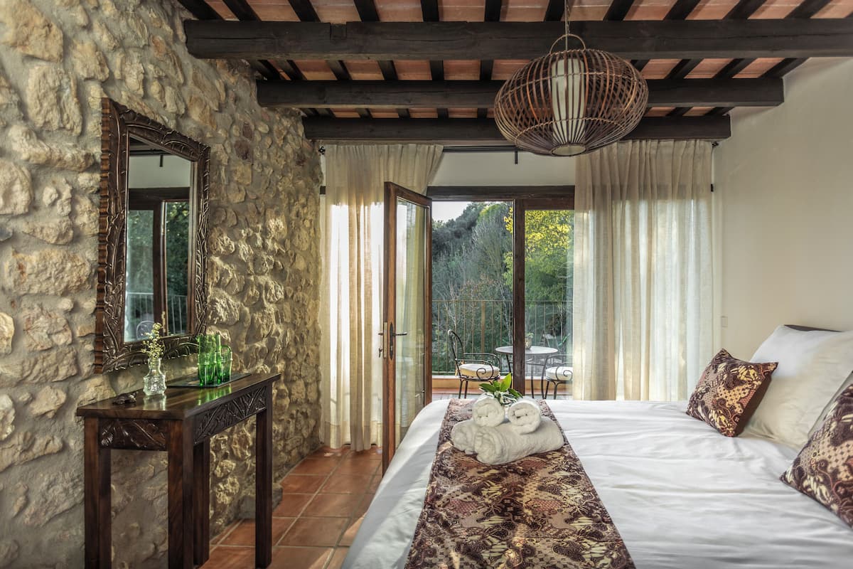 Une chambre avec des murs en pierre, un plafond en bois, un grand lit avec une literie à motifs, une table de chevet avec des lunettes, et une vue sur un jardin par une porte vitrée.