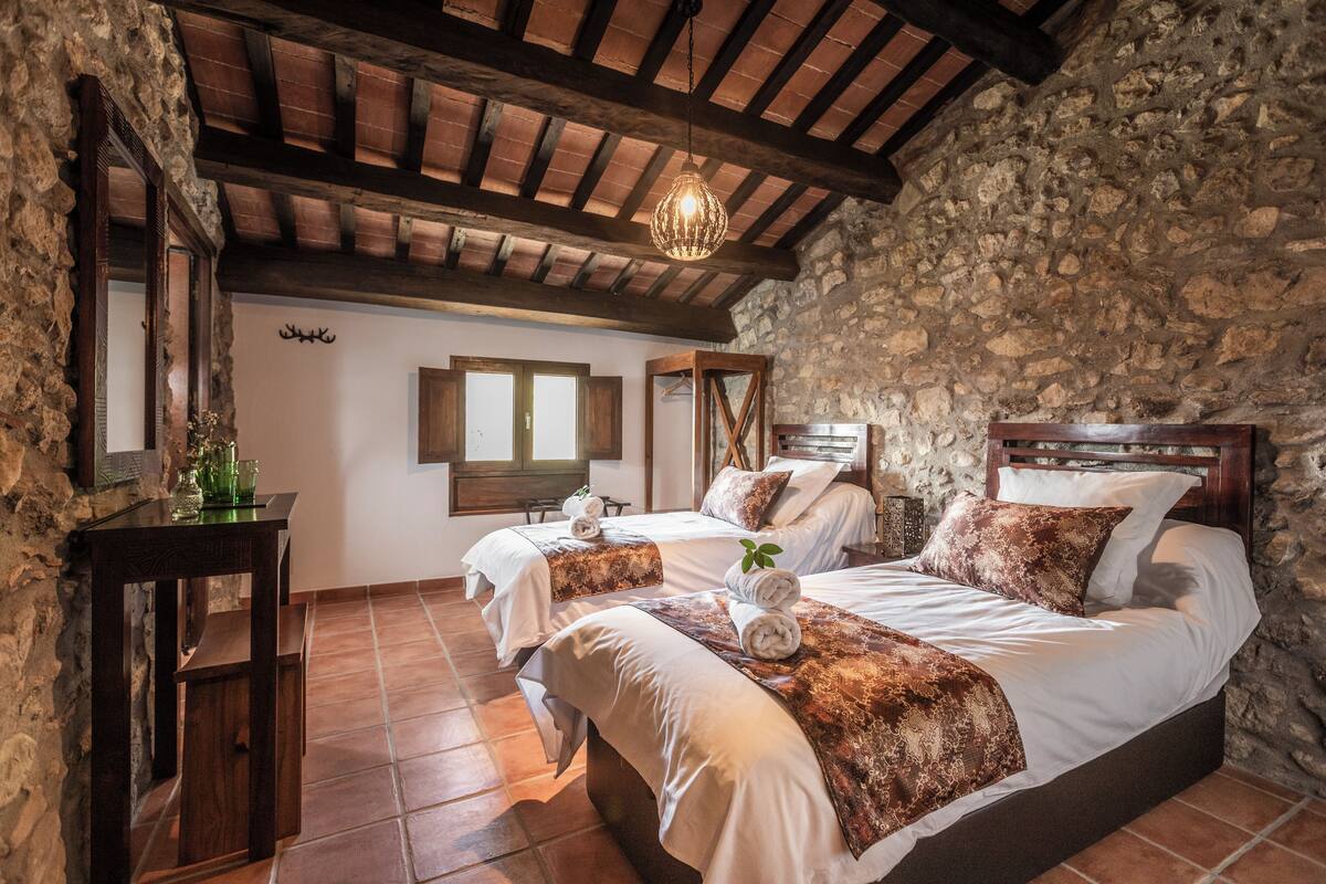 Une chambre à coucher rustique avec deux lits, des murs en pierre, des poutres en bois et un luminaire suspendu.