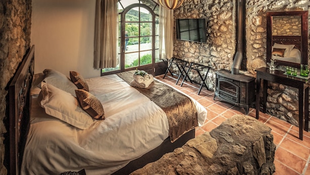 Een rustieke slaapkamer met een stenen muur, een groot bed met veel kussens, een houten hoofdbord, een stenen bank, een open haard en een groot raam met gordijnen.
