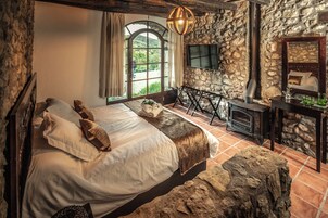 Un dormitorio rústico con pared de piedra, una cama grande con muchas almohadas, cabecero de madera, un banco de piedra, chimenea y una ventana grande con cortinas.