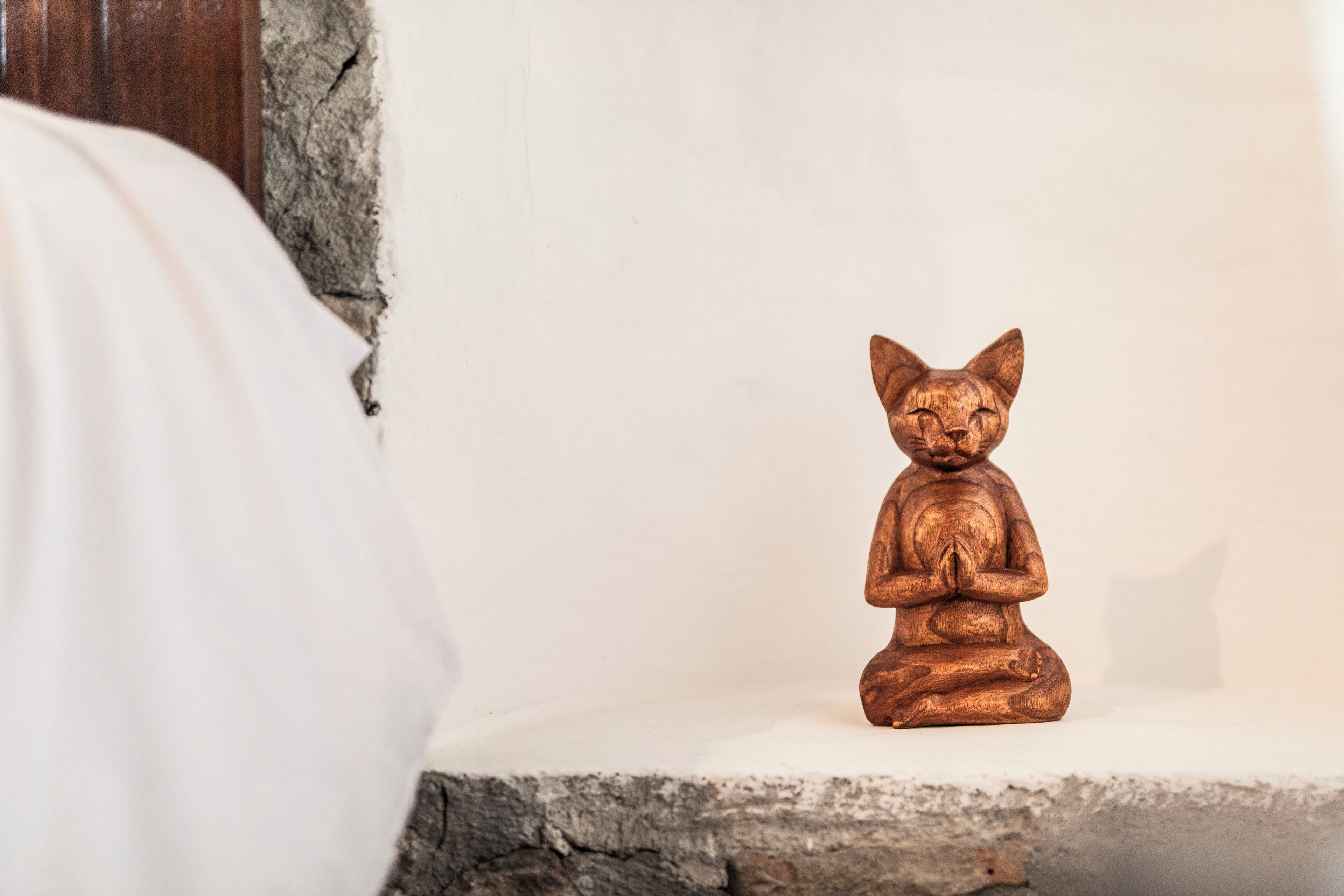 A small wooden statue of a cat sitting on a white shelf against a plain wall.