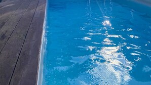 Una piscina rectangular con un deck de madera que la rodea. Hay dos escaleras metálicas para entrar y salir. Un banco de metal está apoyado contra la pared. La piscina está bordeada por una valla blanca y hay un muro de concreto detrás.
