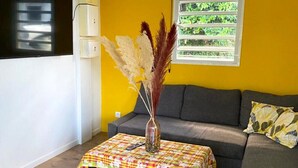A living room with a sofa, a coffee table with a patterned tablecloth, a vase with dried plants, and a window with a view of greenery.