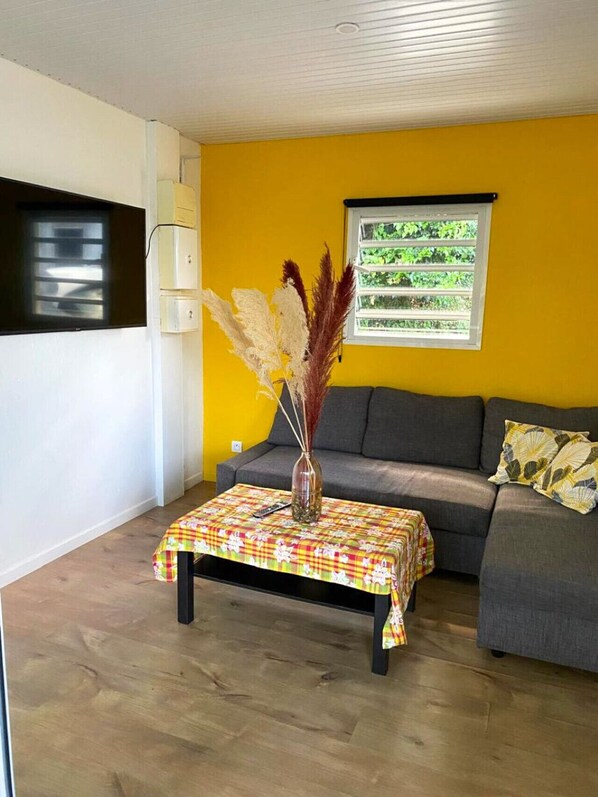 A living room with a sofa, a coffee table with a patterned tablecloth, a vase with dried plants, and a window with a view of greenery.