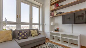 Uma sala de estar com um sofá cinzento, almofadas estampadas, um tapete com padrão, uma estante com livros e objetos decorativos, e uma TV em um suporte branco.